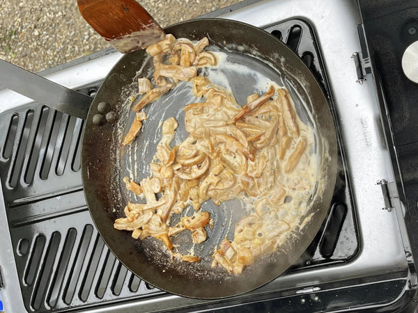 A top-down view of a portable gas stove with a frying pan containing sautéed onions and cream sauce with Russula mushrooms, with a wooden spatula.