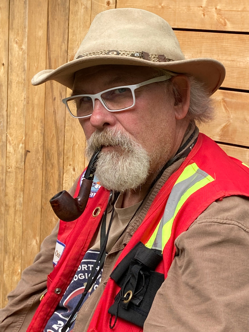 Martin Osis wearing a beige hat, glasses, and a red safety vest with a pipe in front of a wooden background.