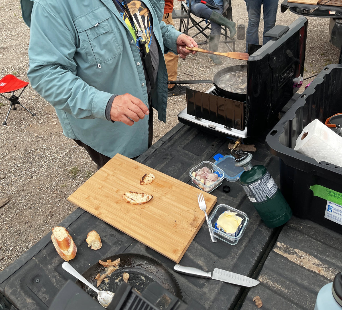 Martin Osis cooks foraged wild mushrooms at an outdoor campsite next to a black portable stove, with a wooden cutting board, bread, and cooking containers on a truck bed.